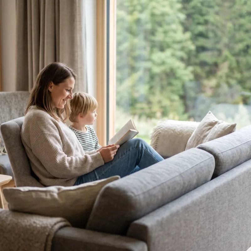 Mutter und Kind lesen gemütlich auf dem Sofa mit Waldblick während des Familienurlaubs im Harz.